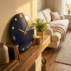 A navy blue wall clock with gold Roman numerals and hands. The clock is round and mounted on a light-colored wall. The clock is next to a wooden shelf with a fern, a candle, and books.