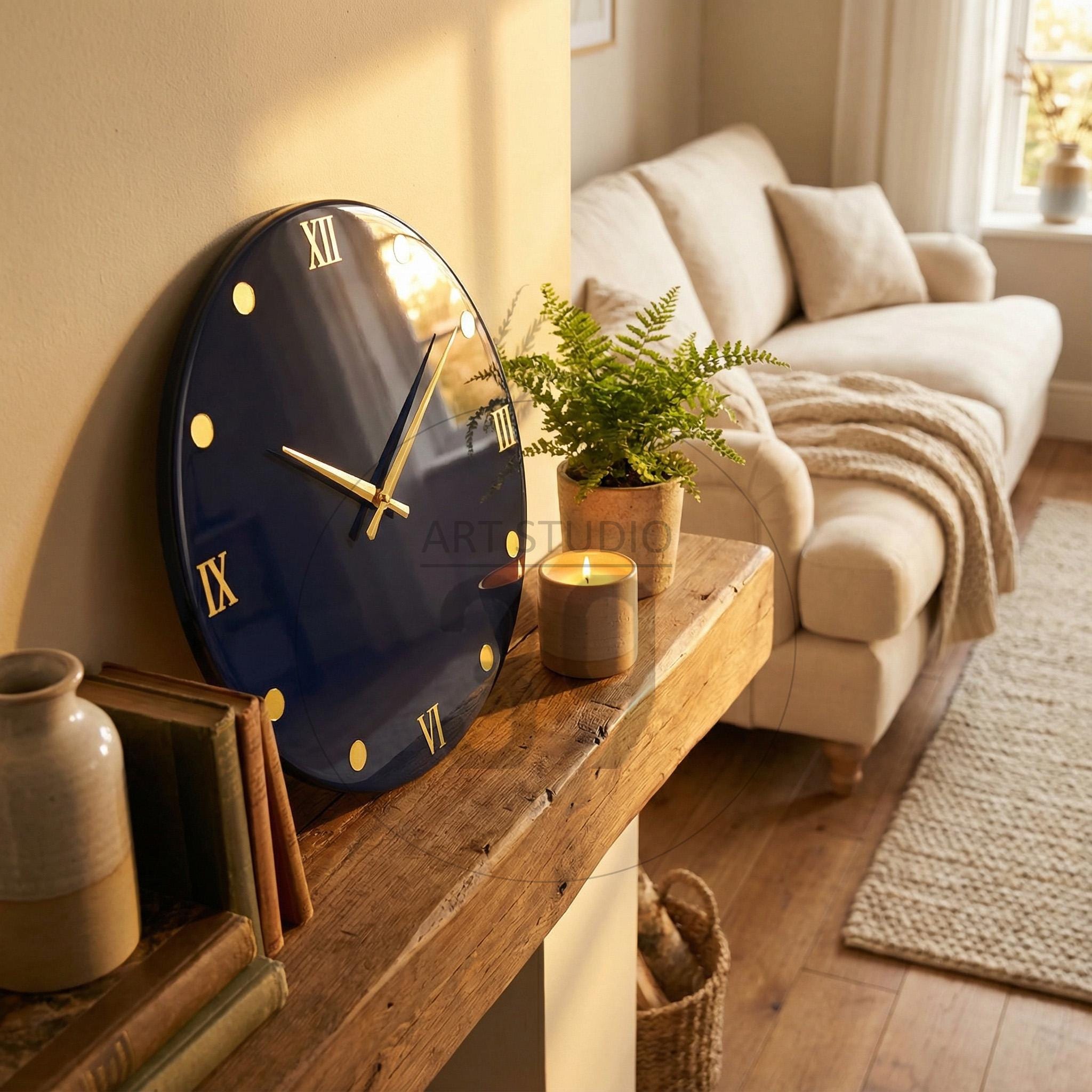 A navy blue wall clock with gold Roman numerals and hands. The clock is round and mounted on a light-colored wall. The clock is next to a wooden shelf with a fern, a candle, and books.