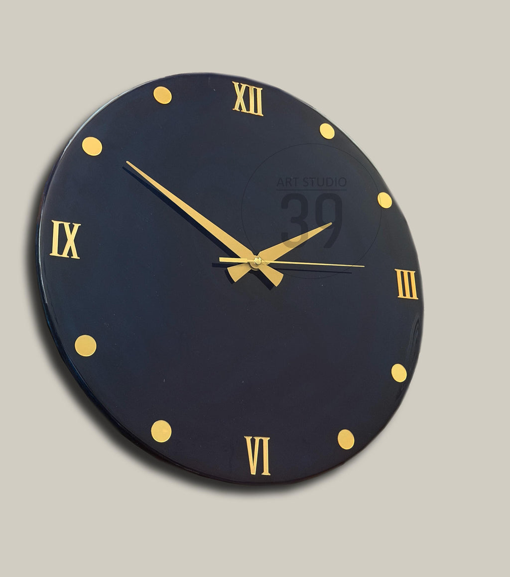 A round, black wall clock with gold Roman numerals and hands. Gold dots mark the hours. The clock face has the text "ART STUDIO 39".