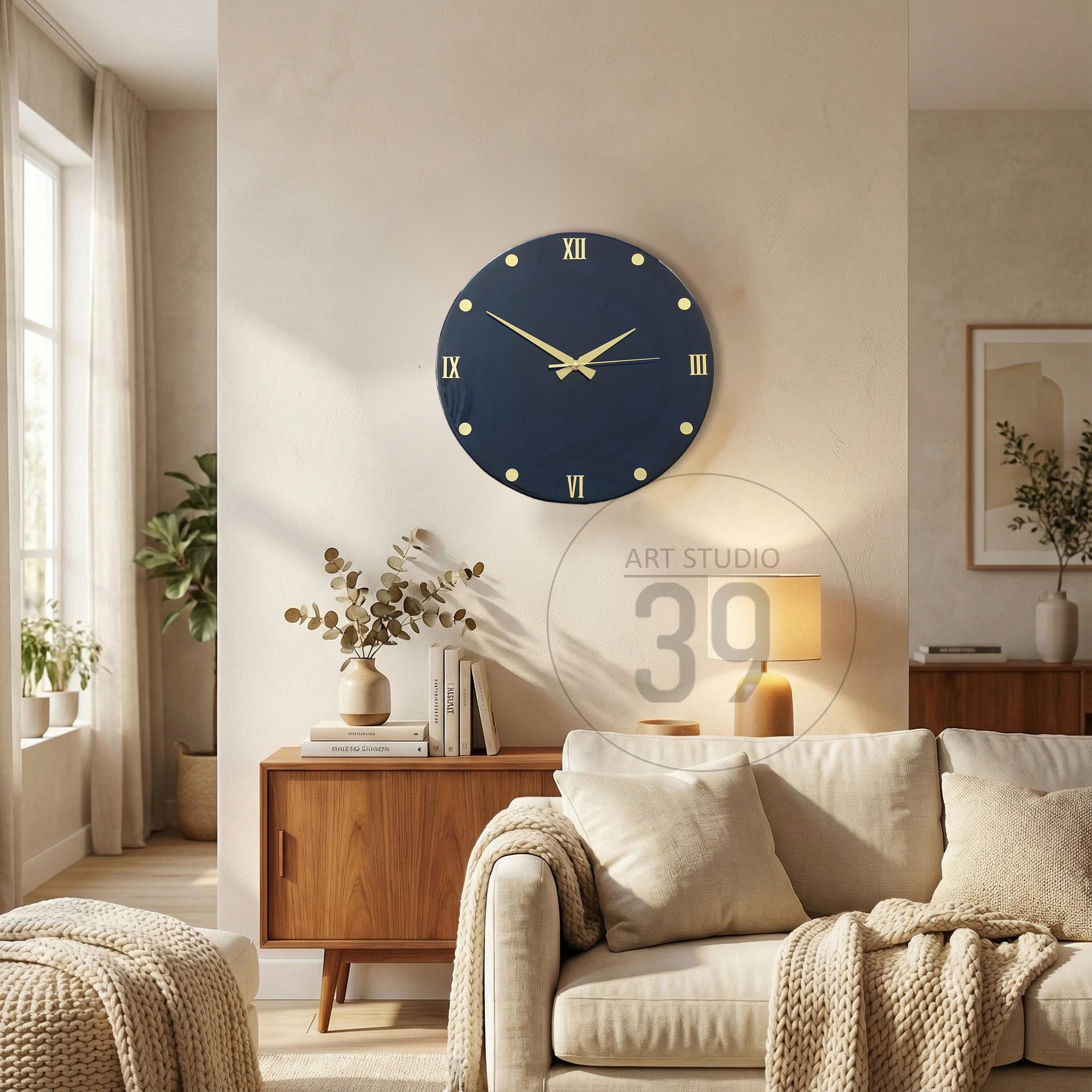 A navy blue wall clock with gold-colored hands and Roman numerals. The clock is round and has gold-colored dots marking the hours. The clock is hanging on a light-colored wall in a living room setting.