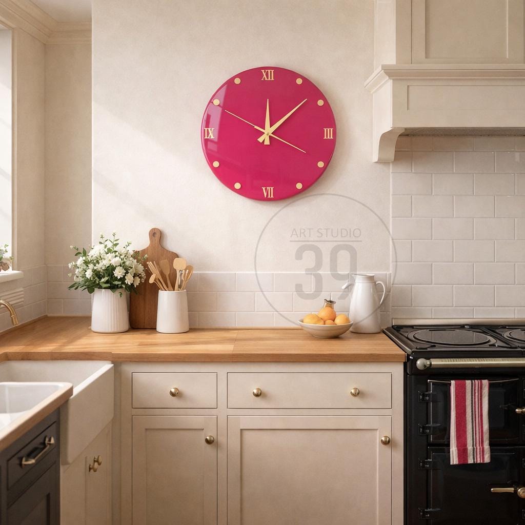 A vibrant magenta wall clock with gold-colored hands and Roman numerals. The clock is round and hangs on a light-colored wall in a kitchen setting. The kitchen features white cabinets, a wooden countertop, and a black stove.