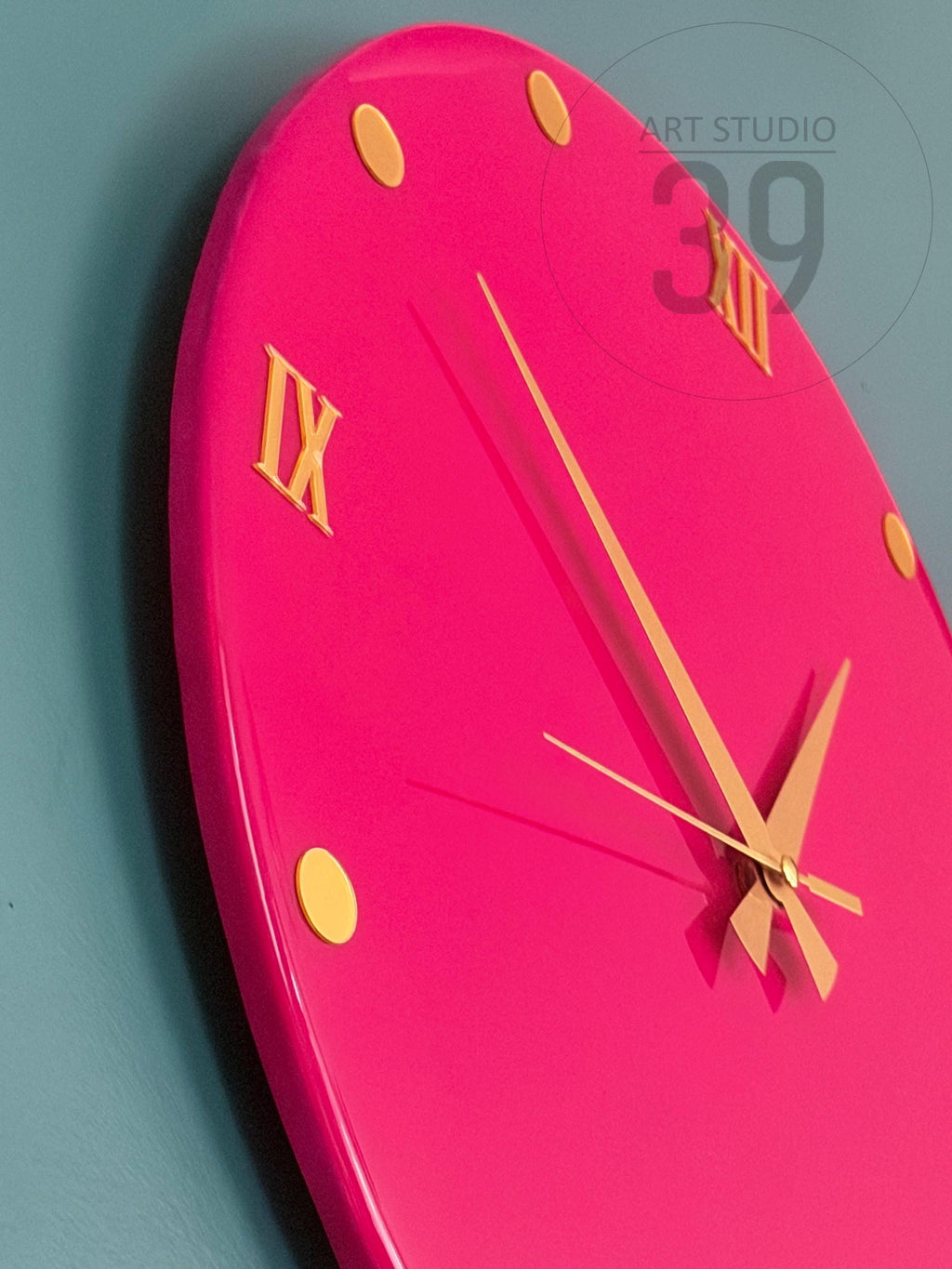 A bright pink, oval-shaped wall clock with gold-colored Roman numerals and hands. The clock has a glossy finish and is accented with small, round gold markers. The text "ART STUDIO 39" is visible on the clock face.
