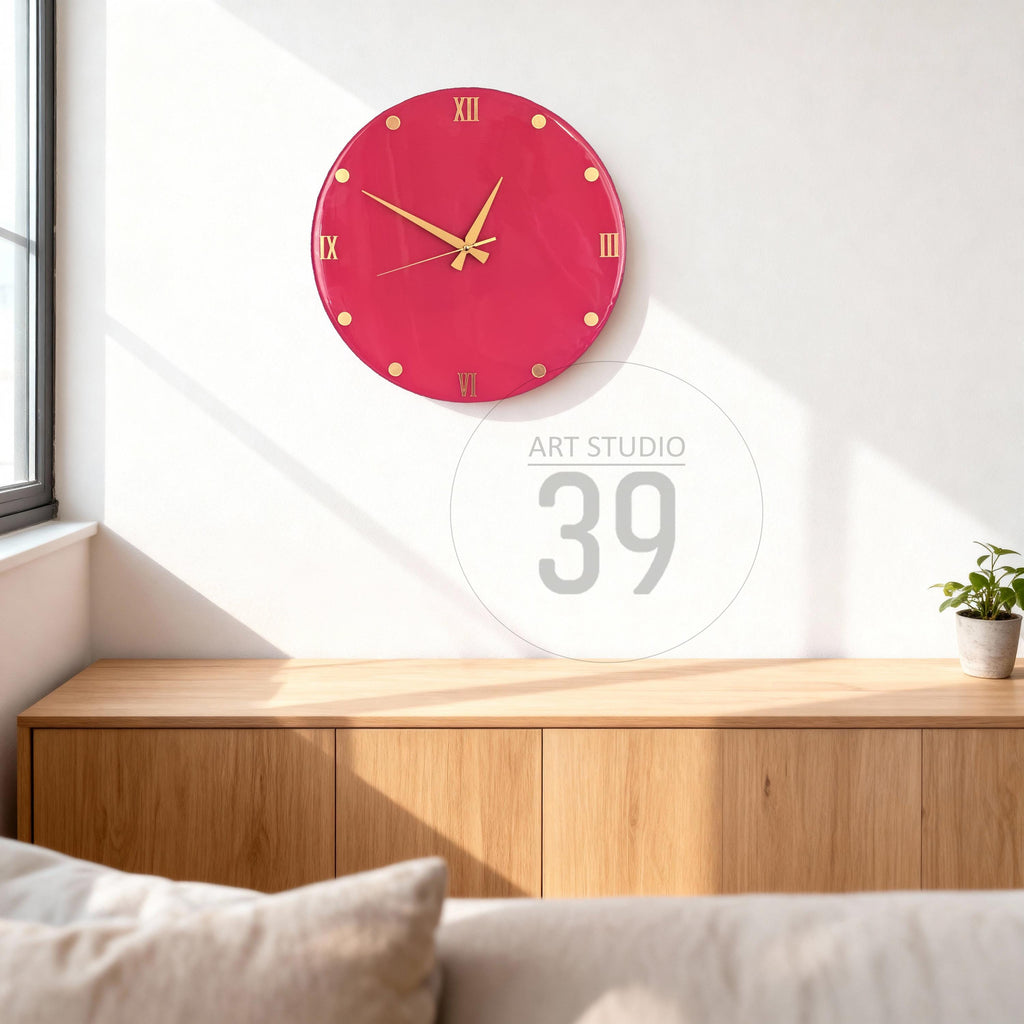 A round, raspberry-colored wall clock with gold-colored hands and hour markers. Roman numerals mark the hours. The clock is mounted on a white wall above a wooden cabinet. The text "ART STUDIO 39" is visible.