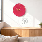 A round, raspberry-colored wall clock with gold-colored hands and hour markers. Roman numerals mark the hours. The clock is mounted on a white wall above a wooden cabinet. The text "ART STUDIO 39" is visible.
