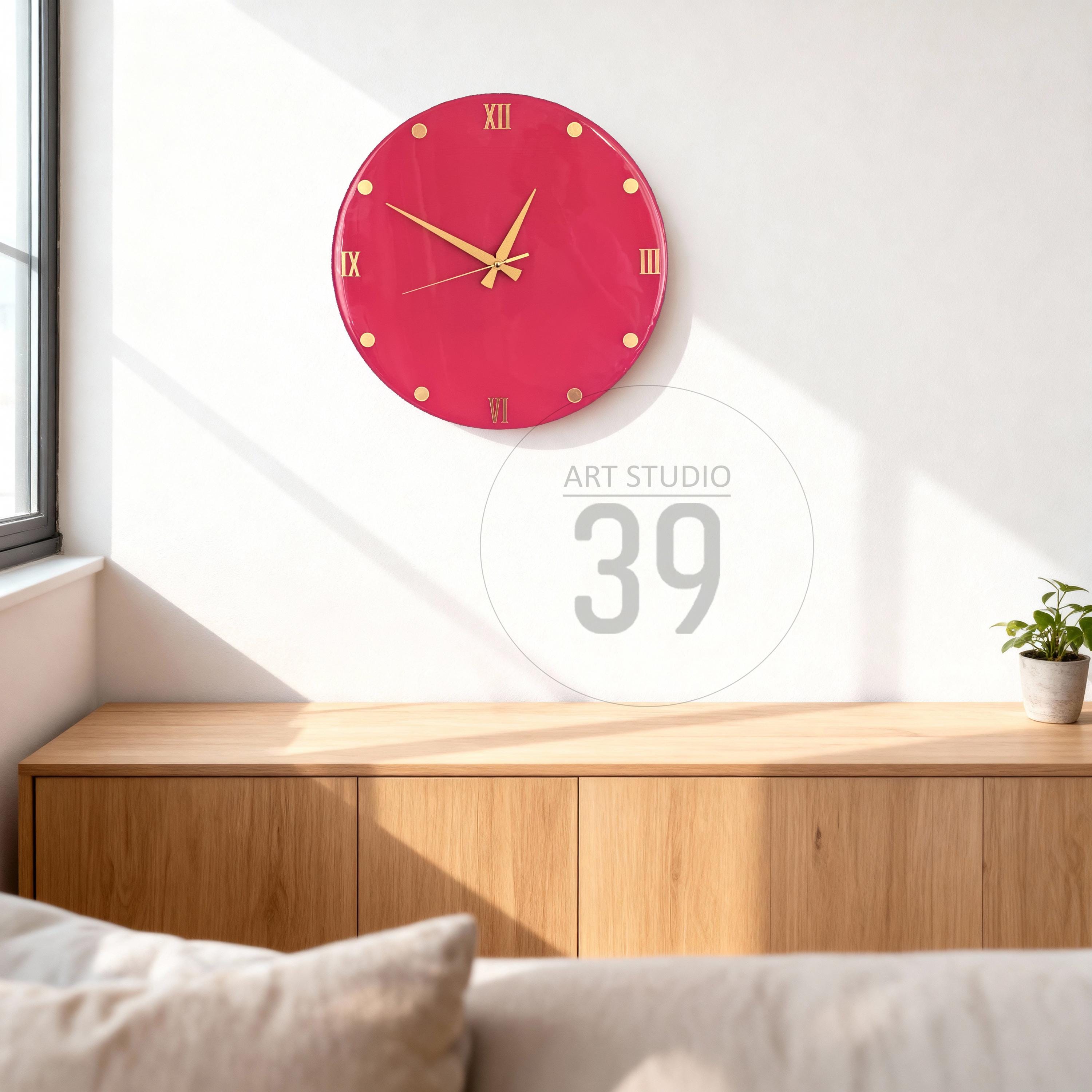 A round, raspberry-colored wall clock with gold-colored hands and hour markers. Roman numerals mark the hours. The clock is mounted on a white wall above a wooden cabinet. The text "ART STUDIO 39" is visible.