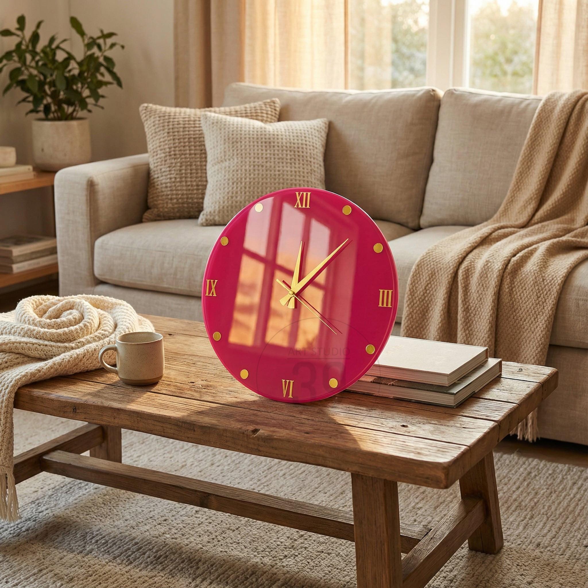 A round, magenta wall clock with gold-colored hands and hour markers. The clock features Roman numerals and is displayed on a rustic wooden coffee table. The clock is approximately 12 inches in diameter.