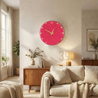 A round, hot pink wall clock with gold-colored hands and Roman numerals. The clock is mounted on a light-colored wall in a living room setting. The clock's diameter is approximately 12 inches.