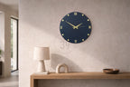 A navy blue wall clock with gold Roman numerals and hands. The clock is round and mounted on a light beige wall. A beige table lamp, a decorative arch, and a wooden bowl are on a wooden shelf below the clock.