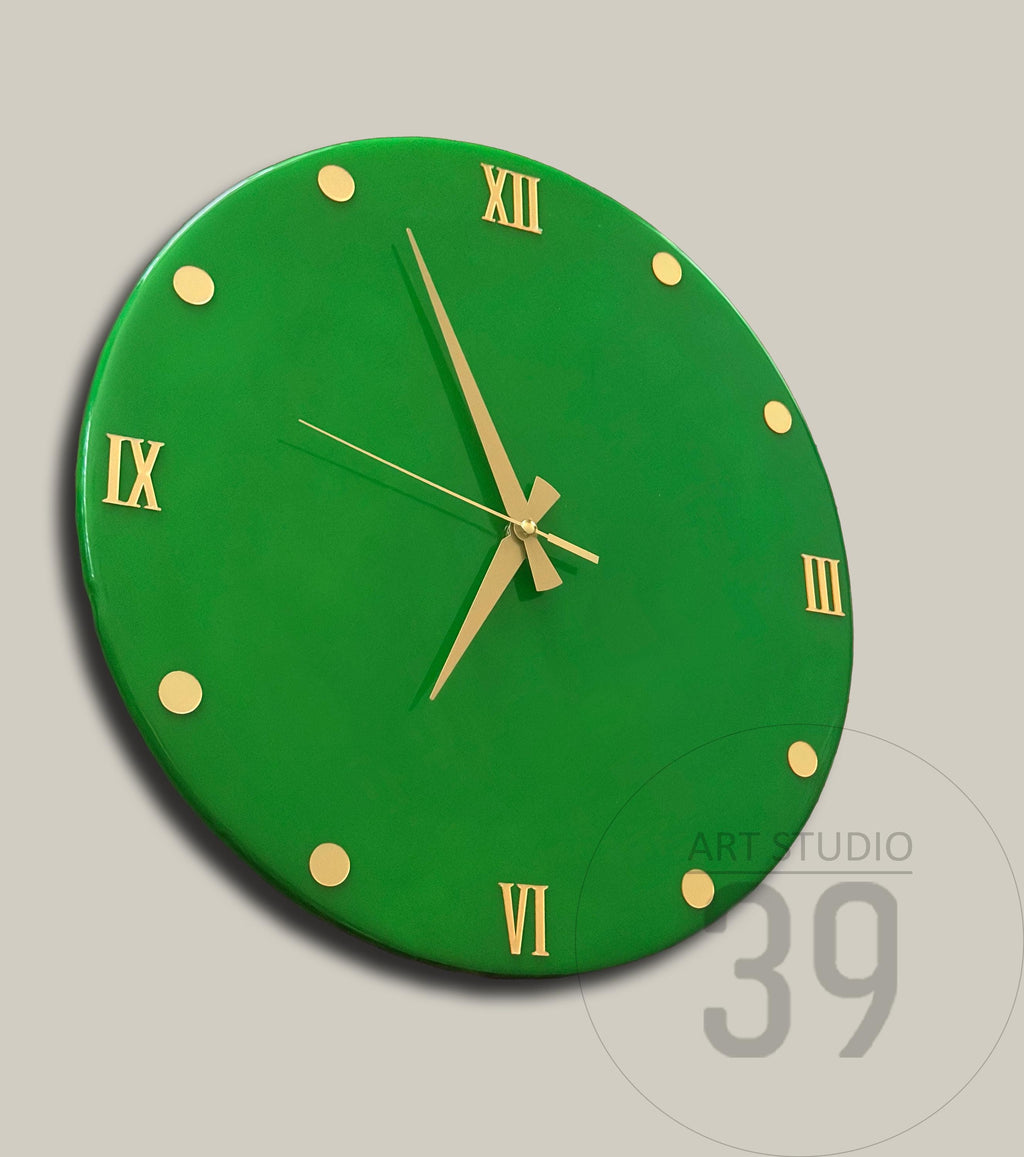 A round, green wall clock with gold-colored Roman numerals and hands. The clock face has gold-colored dots marking the hours. The clock is a modern design.