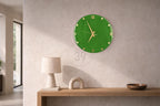 A green wall clock with gold-colored hands and Roman numerals. The clock is round and mounted on a light-colored wall. The clock is a decorative item for a home or office.