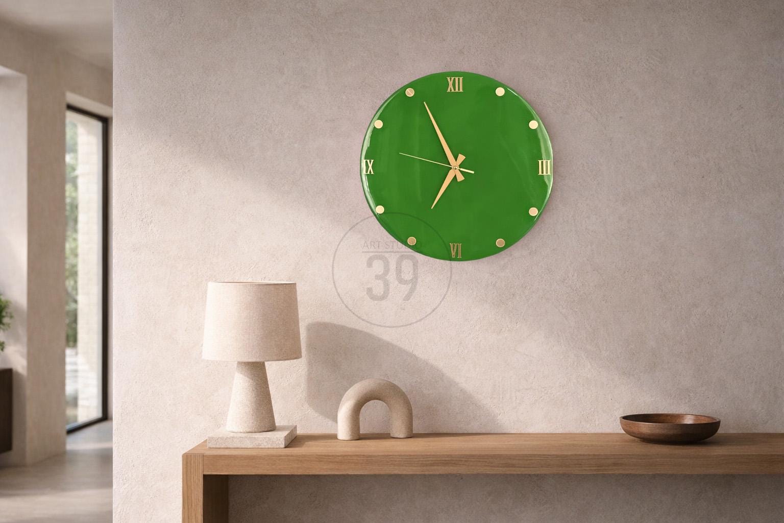 A green wall clock with gold-colored hands and Roman numerals. The clock is round and mounted on a light-colored wall. The clock is a decorative item for a home or office.