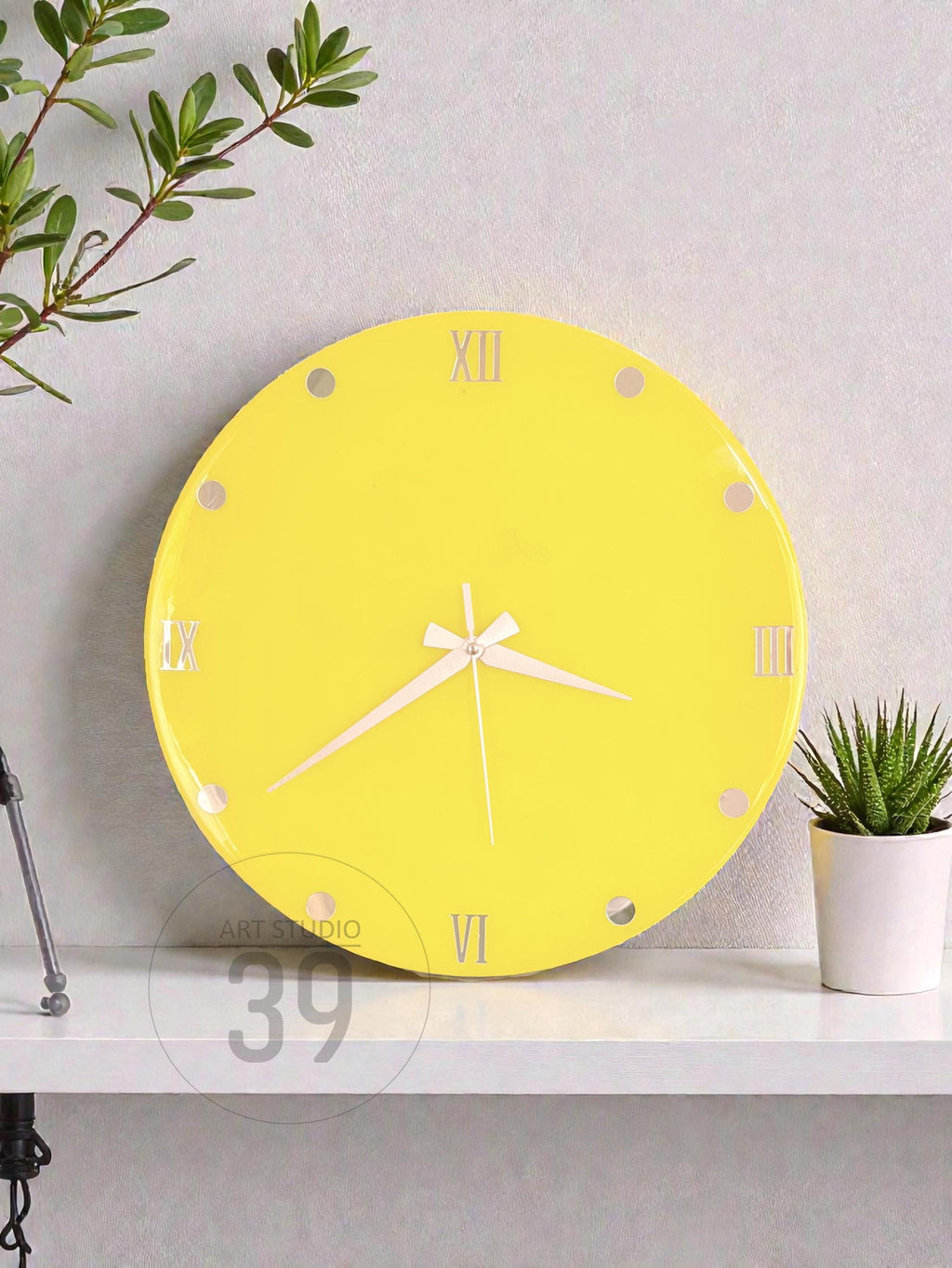 A round, bright yellow wall clock with white hands and Roman numeral hour markers. The clock has a minimalist design and is displayed on a white shelf. A small potted plant is to the right of the clock.