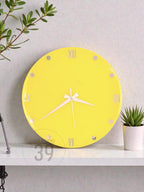 A round, bright yellow wall clock with white hands and Roman numeral hour markers. The clock has a minimalist design and is displayed on a white shelf. A small potted plant is to the right of the clock.