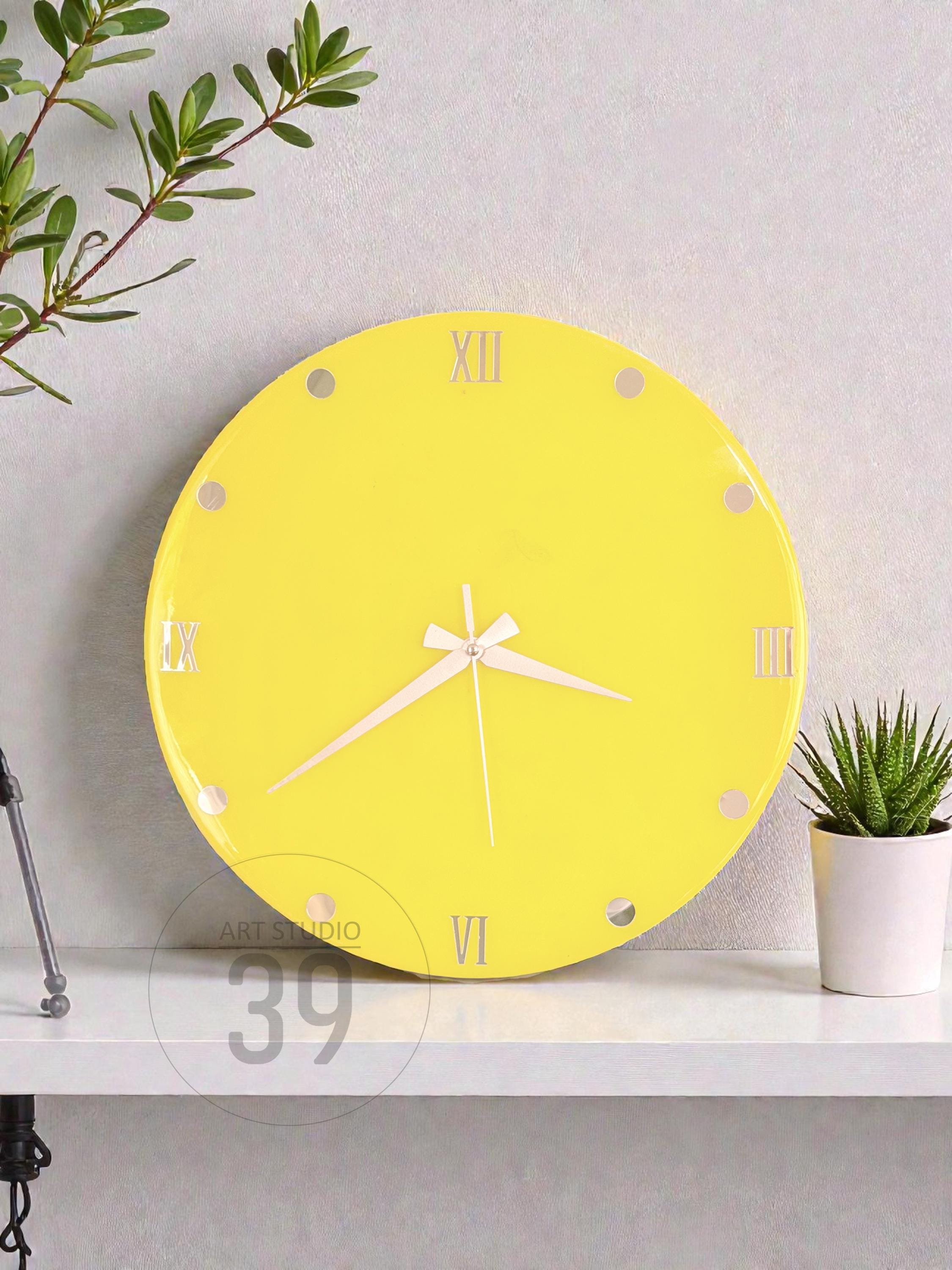 A round, bright yellow wall clock with white hands and Roman numeral hour markers. The clock has a minimalist design and is displayed on a white shelf. A small potted plant is to the right of the clock.