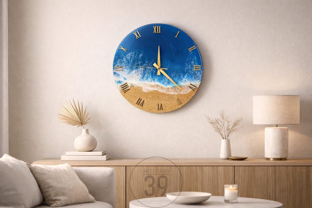 Round wall clock with a beach scene design. The clock face features a blue ocean, white waves, and a sandy beach. Gold Roman numerals and clock hands complete the design. The clock is mounted on a white wall.