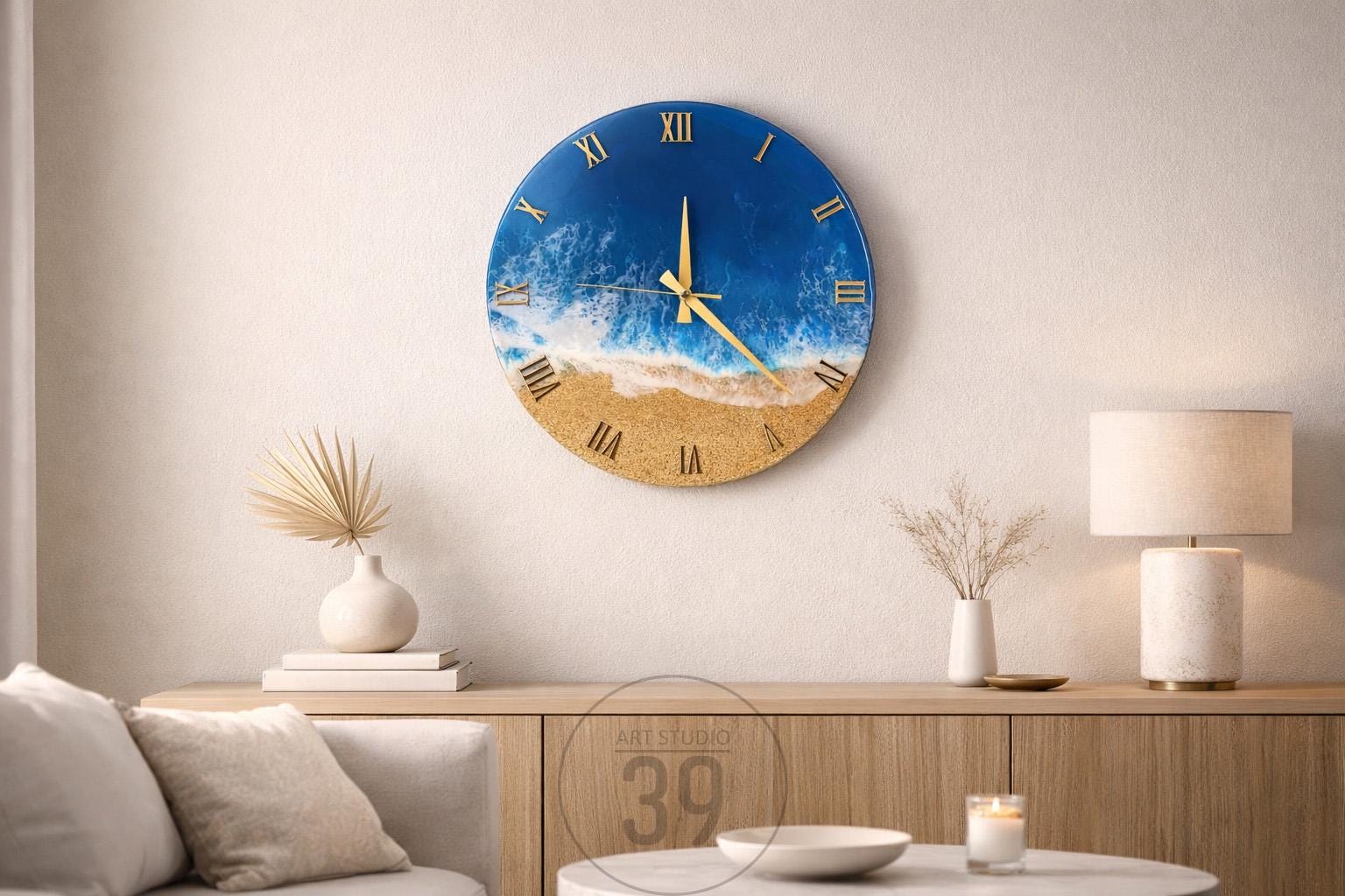 Round wall clock with a beach scene design. The clock face features a blue ocean, white waves, and a sandy beach. Gold Roman numerals and clock hands complete the design. The clock is mounted on a white wall.
