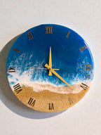 Round wall clock with a beach scene design. The clock face features a blue ocean, white waves, and a sandy beach. Gold Roman numerals mark the hours, and gold hands indicate the time. The clock has a diameter of approximately 12 inches.