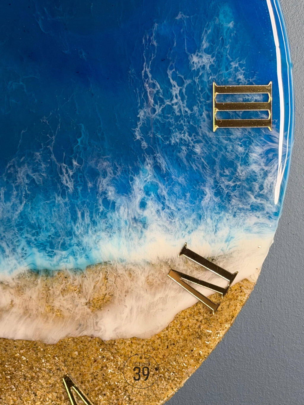 A round wall clock with a beach-themed design. The clock face features a blue ocean with white foamy waves, a sandy beach, and gold Roman numerals. The number 39 is visible on the clock face.