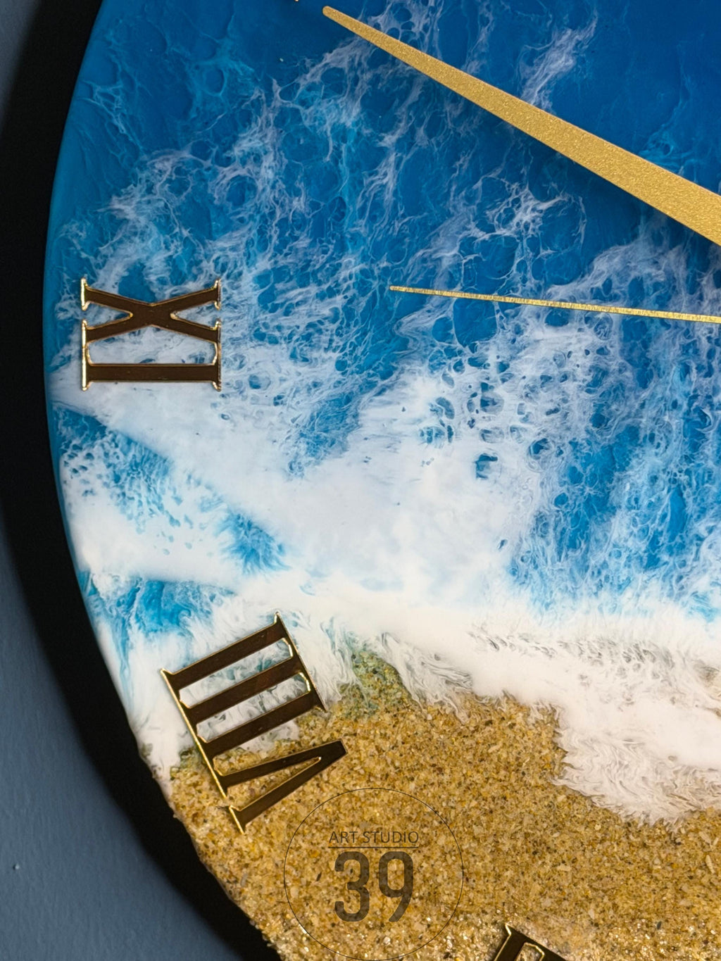 Round wall clock with a beach-themed design. The clock face features a blue and white ocean wave pattern, gold Roman numerals, and gold clock hands. The bottom of the clock face has a sandy beach texture.