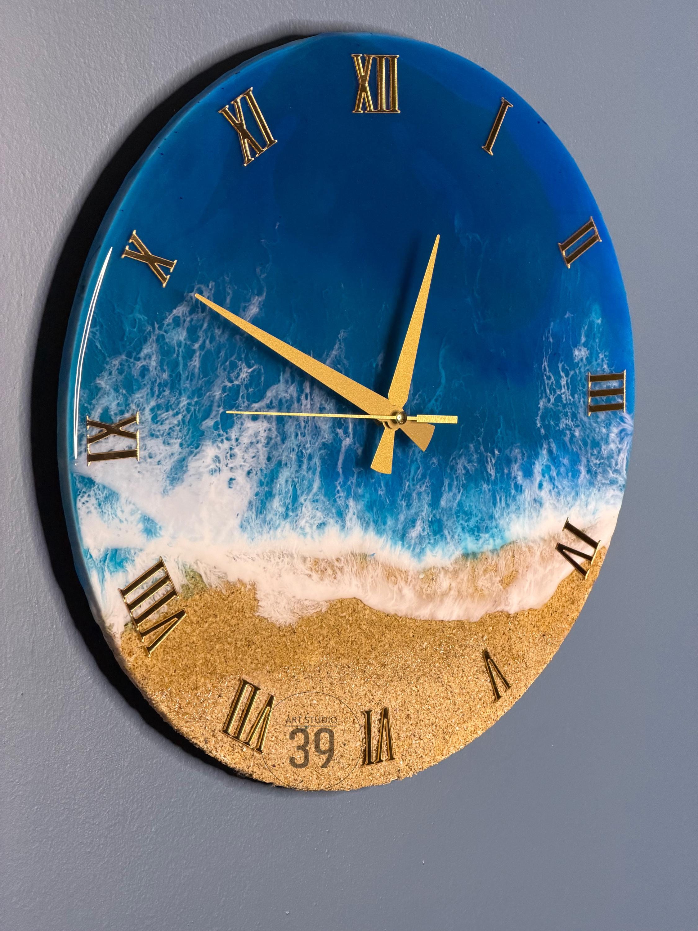 A round wall clock with a beach-themed design. The top half features a deep blue ocean with white foamy waves, transitioning to a sandy beach at the bottom. Gold Roman numerals mark the hours, and gold hands indicate the time.