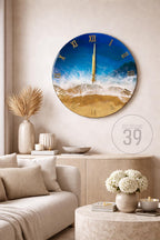 A round wall clock with a beach scene design. The clock face features a blue sky, white waves, and a golden sand beach. Gold Roman numerals mark the hours. The clock is displayed in a living room setting.