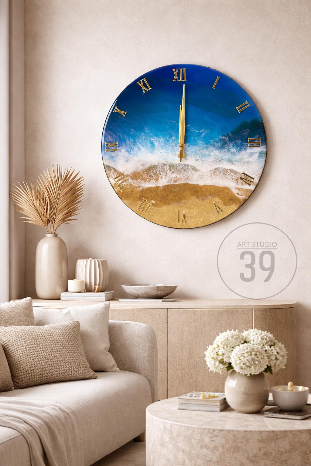A round wall clock with a beach scene design. The clock face features a blue sky, white waves, and a golden sand beach. Gold Roman numerals mark the hours. The clock is displayed in a living room setting.