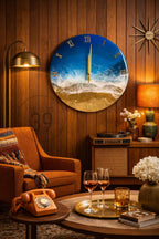 A round wall clock with a beach scene design. The clock face features a blue ocean, white waves, and a golden sand beach. Gold Roman numerals mark the hours. The clock is mounted on a wood-paneled wall.