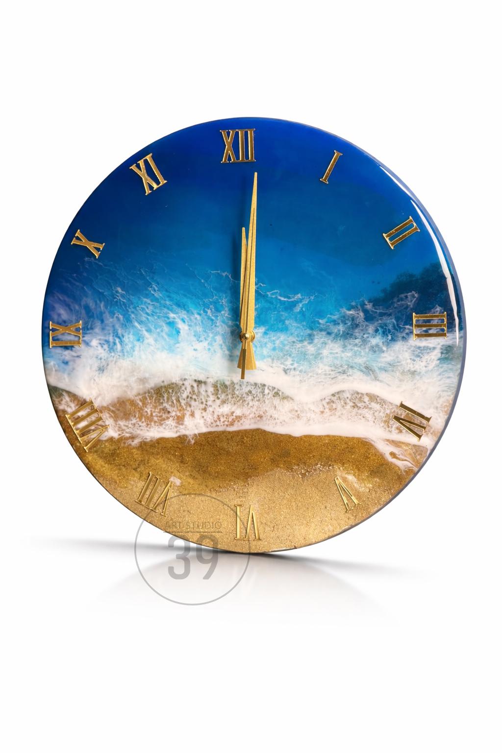 Round wall clock with a beach scene design. The clock face features a blue ocean with white waves and a golden sand beach. Gold Roman numerals mark the hours, and gold hands indicate the time.