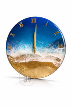 Round wall clock with a beach scene design. The clock face features a blue ocean with white waves and a golden sand beach. Gold Roman numerals mark the hours, and gold hands indicate the time.