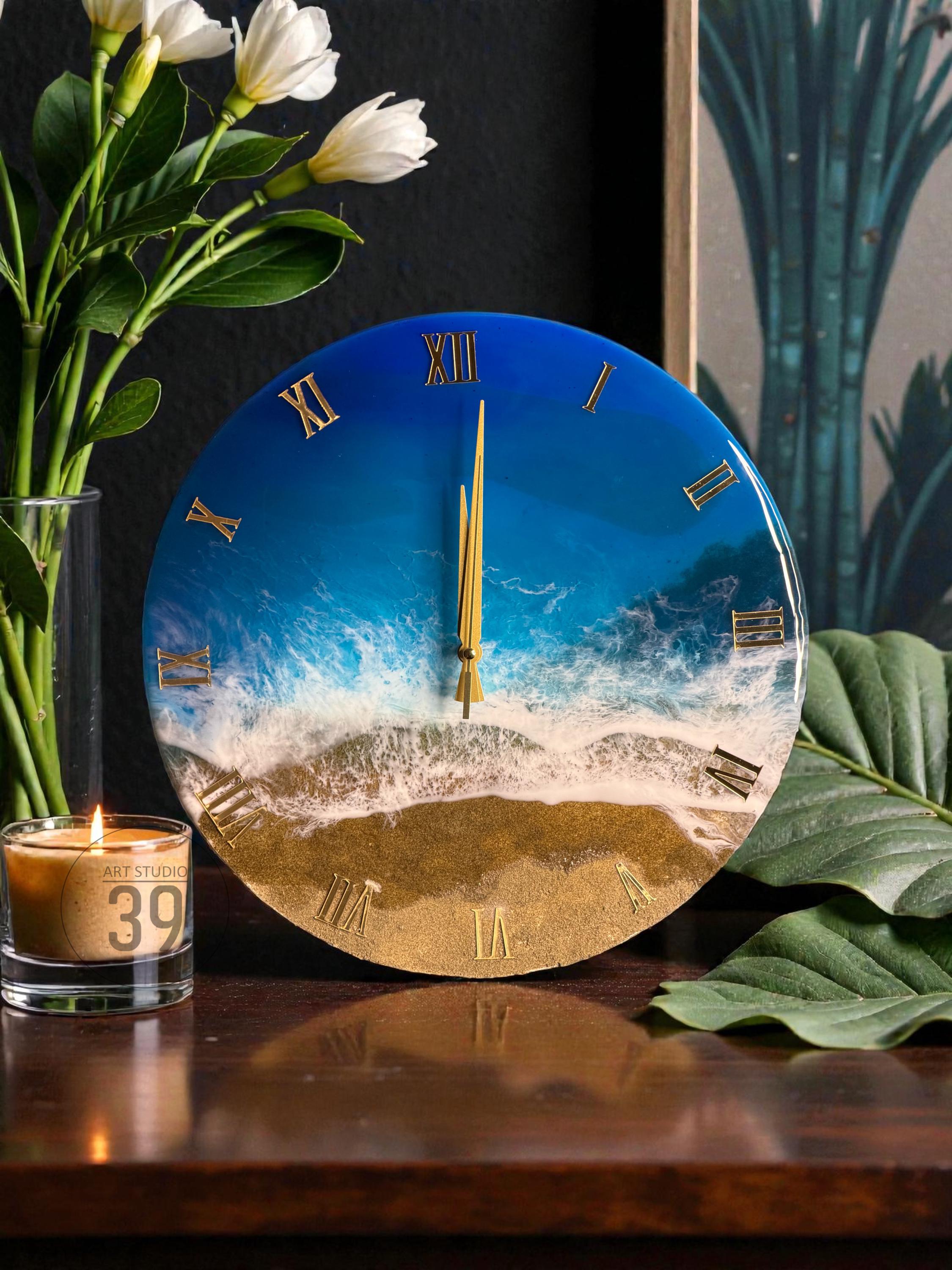 Round wall clock with a beach scene design. The clock face features a blue ocean, white waves, and golden sand, with gold Roman numerals and clock hands. The clock is a decorative home decor item.
