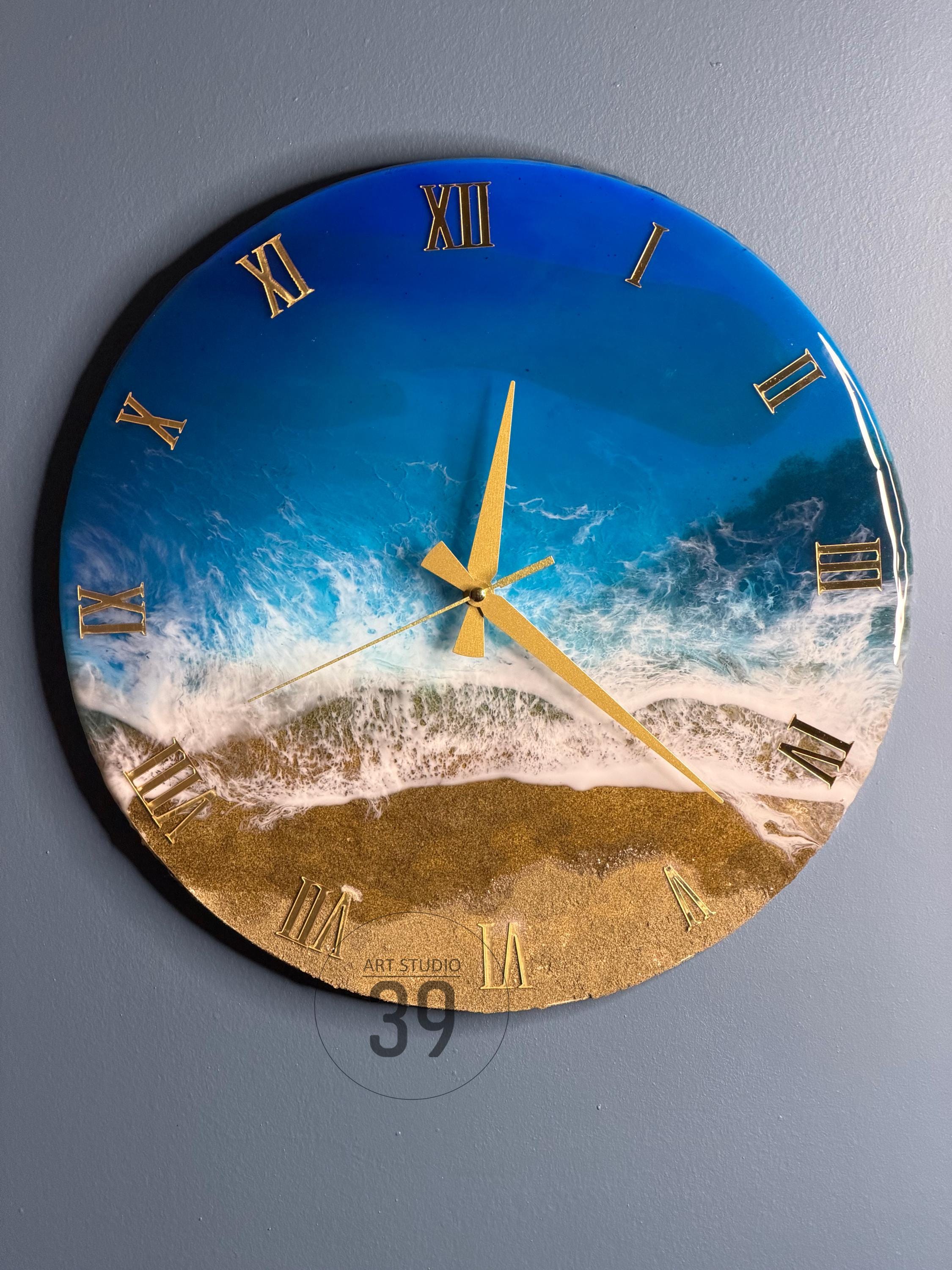 Round wall clock with a beach-themed design. The clock face features a blue ocean, white waves, and a golden sand beach. Gold Roman numerals mark the hours, and gold hands indicate the time.