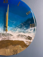 A round wall clock with a beach-themed design. The clock face features a blue ocean with white waves, golden sand, and gold Roman numerals. The clock hands are gold. The text "ART STUDIO 39" is visible.