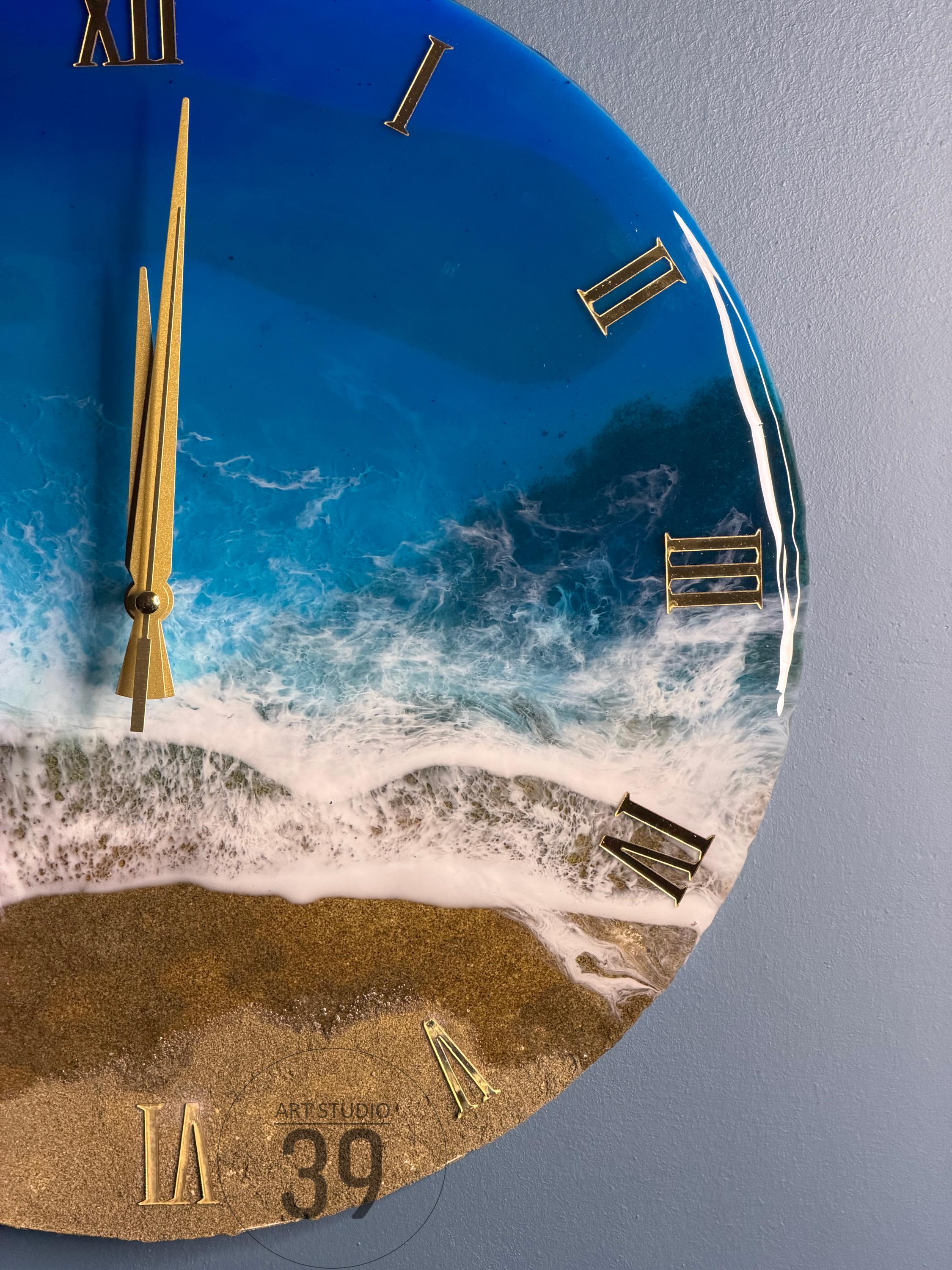 A round wall clock with a beach-themed design. The clock face features a blue ocean with white waves, golden sand, and gold Roman numerals. The clock hands are gold. The text "ART STUDIO 39" is visible.