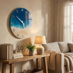 A round wall clock with a beach-themed design. The clock face features a gradient of blue and white, resembling the ocean and sand. Silver Roman numerals mark the hours. The clock is mounted on a beige wall.