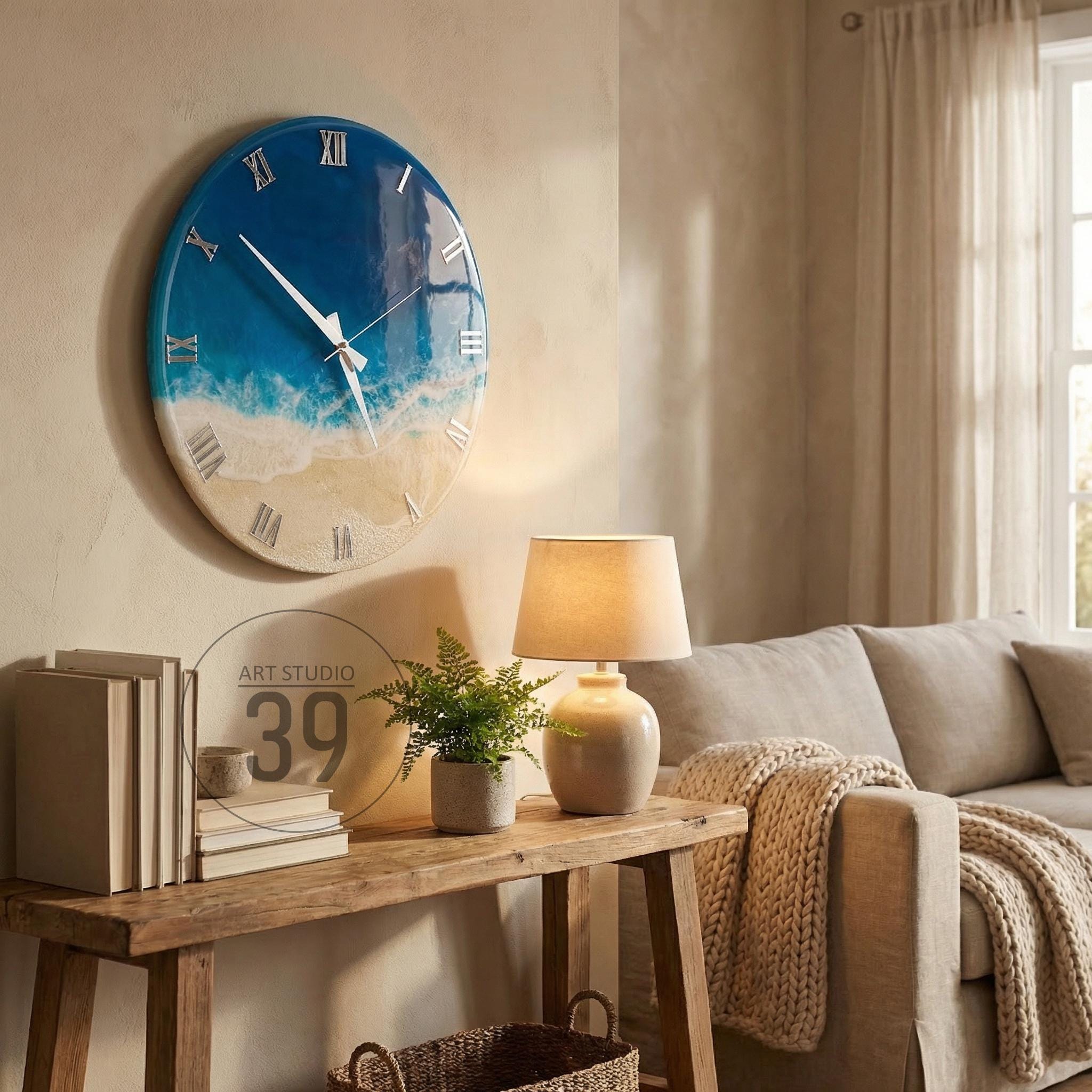 A round wall clock with a beach-themed design. The clock face features a gradient of blue and white, resembling the ocean and sand. Silver Roman numerals mark the hours. The clock is mounted on a beige wall.