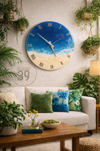 A large, round wall clock with a beach-themed design. The clock face features a blue and white ocean scene with Roman numerals. The clock is mounted on a light-colored wall, surrounded by plants and a sofa with pillows.