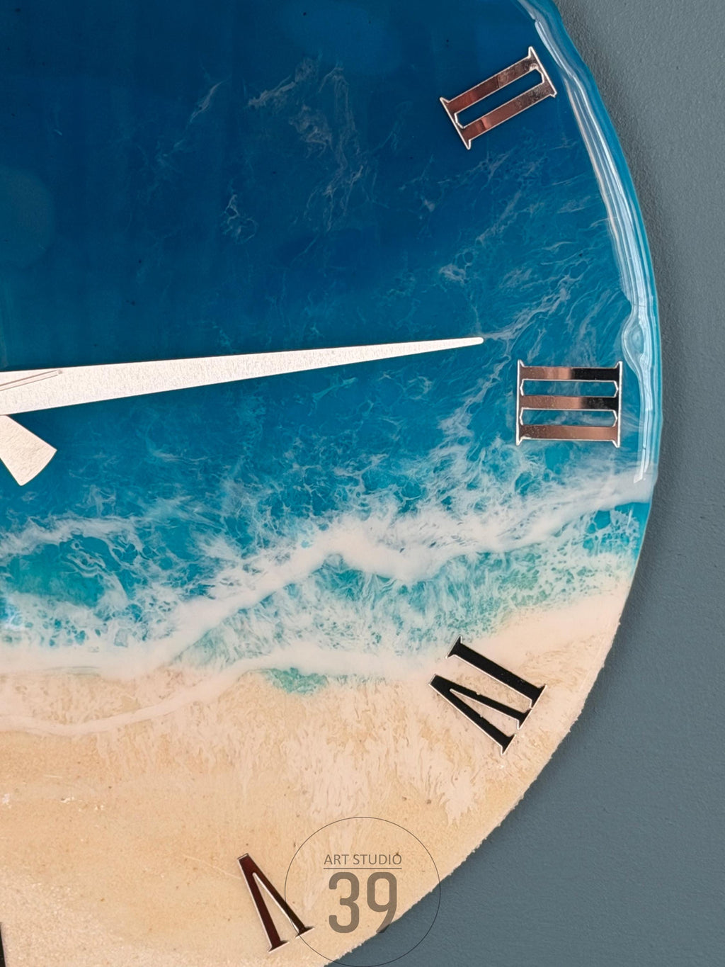 A round wall clock with a beach-themed design. The clock face features a blue ocean, white waves, and a sandy beach. Silver Roman numerals mark the hours. The clock hands are white. The text "ART STUDIO 39" is visible.