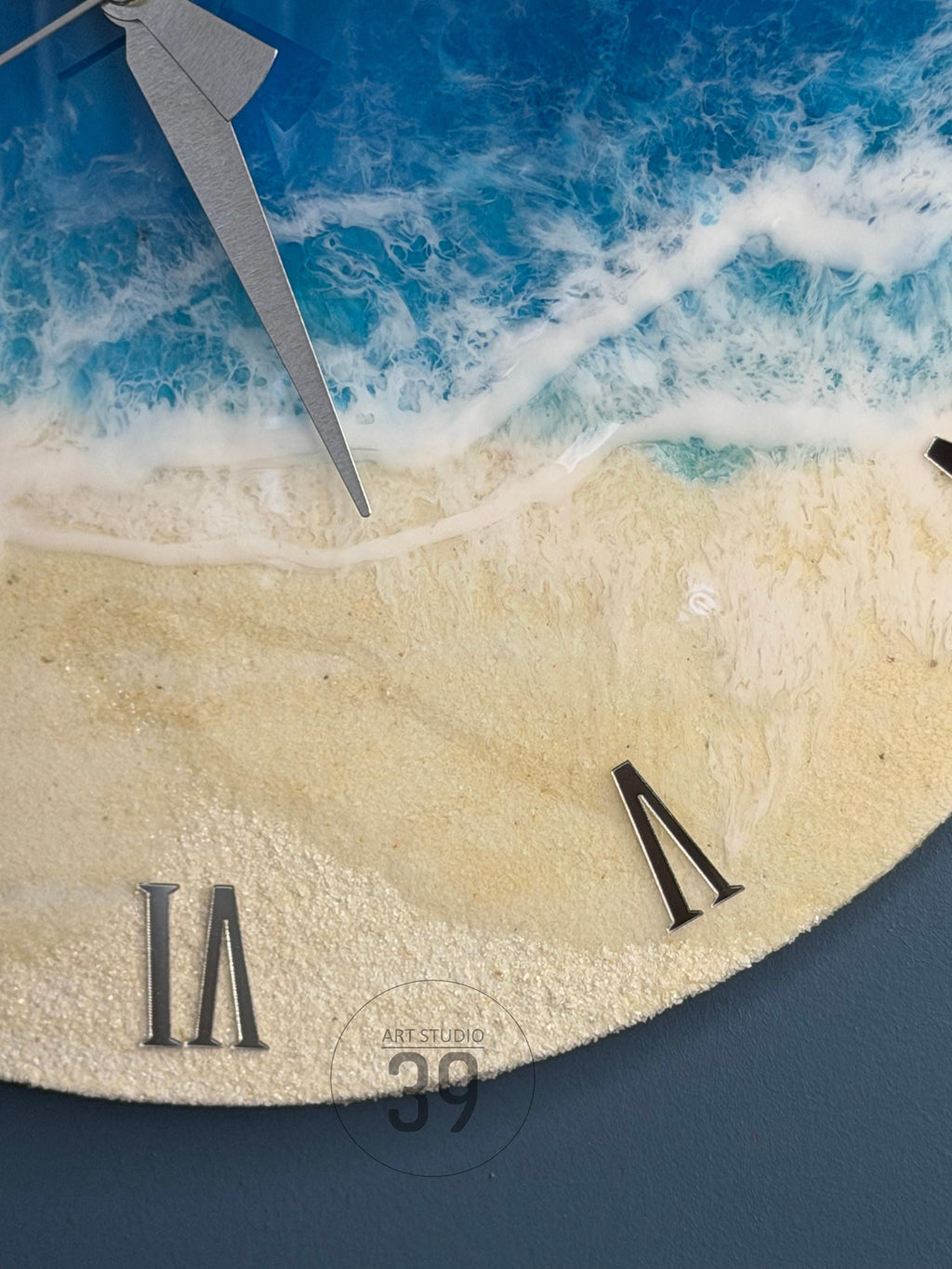 A round wall clock with a beach-themed design. The clock face features a depiction of ocean waves and sandy shore. Silver clock hands and Roman numerals mark the time. The clock has the text "ART STUDIO 39".