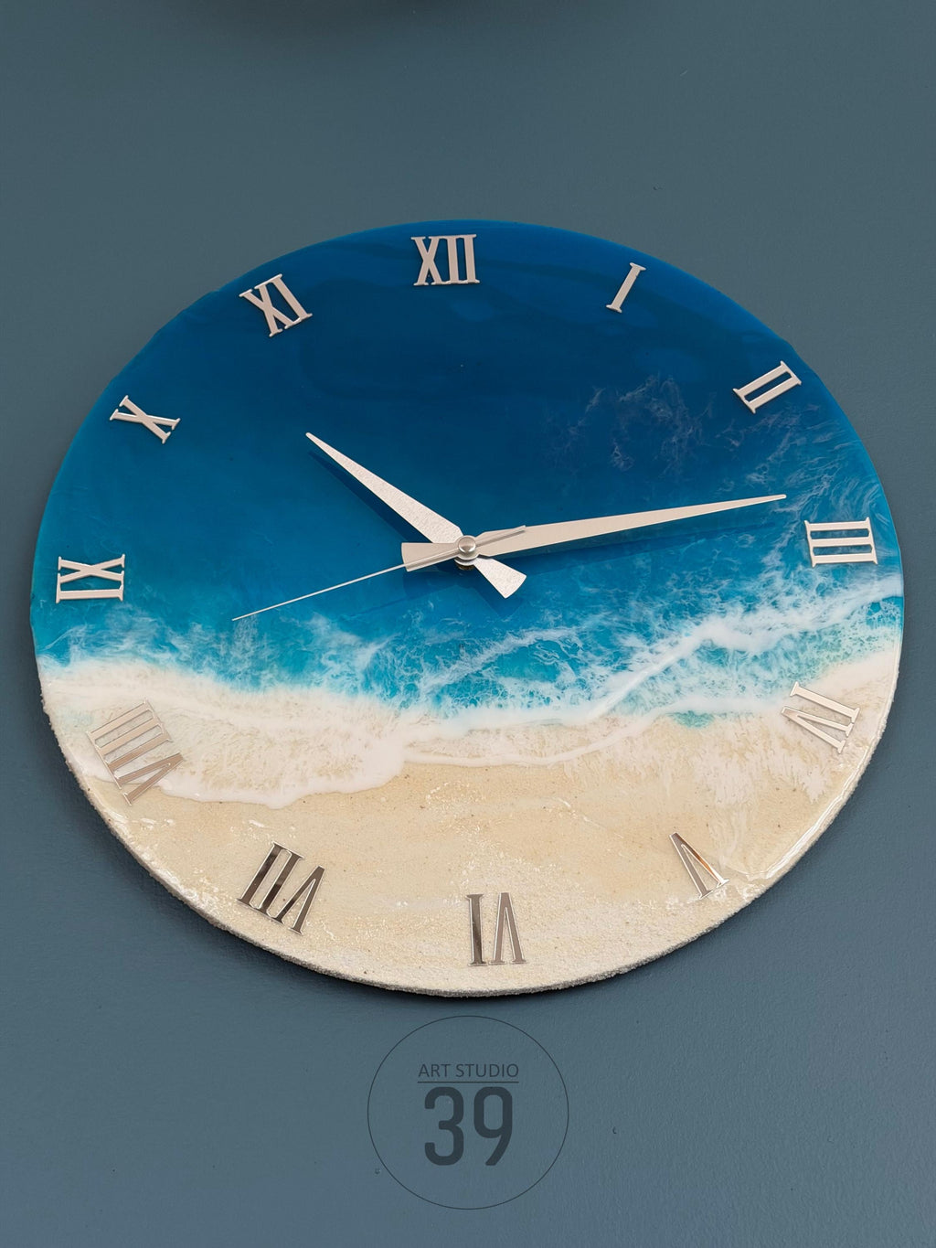 Round wall clock with a beach-themed design. The clock face features a blue and white ocean scene with silver Roman numerals. The clock hands are silver, and the bottom of the clock has the text "ART STUDIO 39".