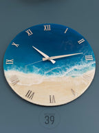 Round wall clock with a beach-themed design. The clock face features a blue and white ocean scene with silver Roman numerals. The clock hands are silver, and the bottom of the clock has the text "ART STUDIO 39".