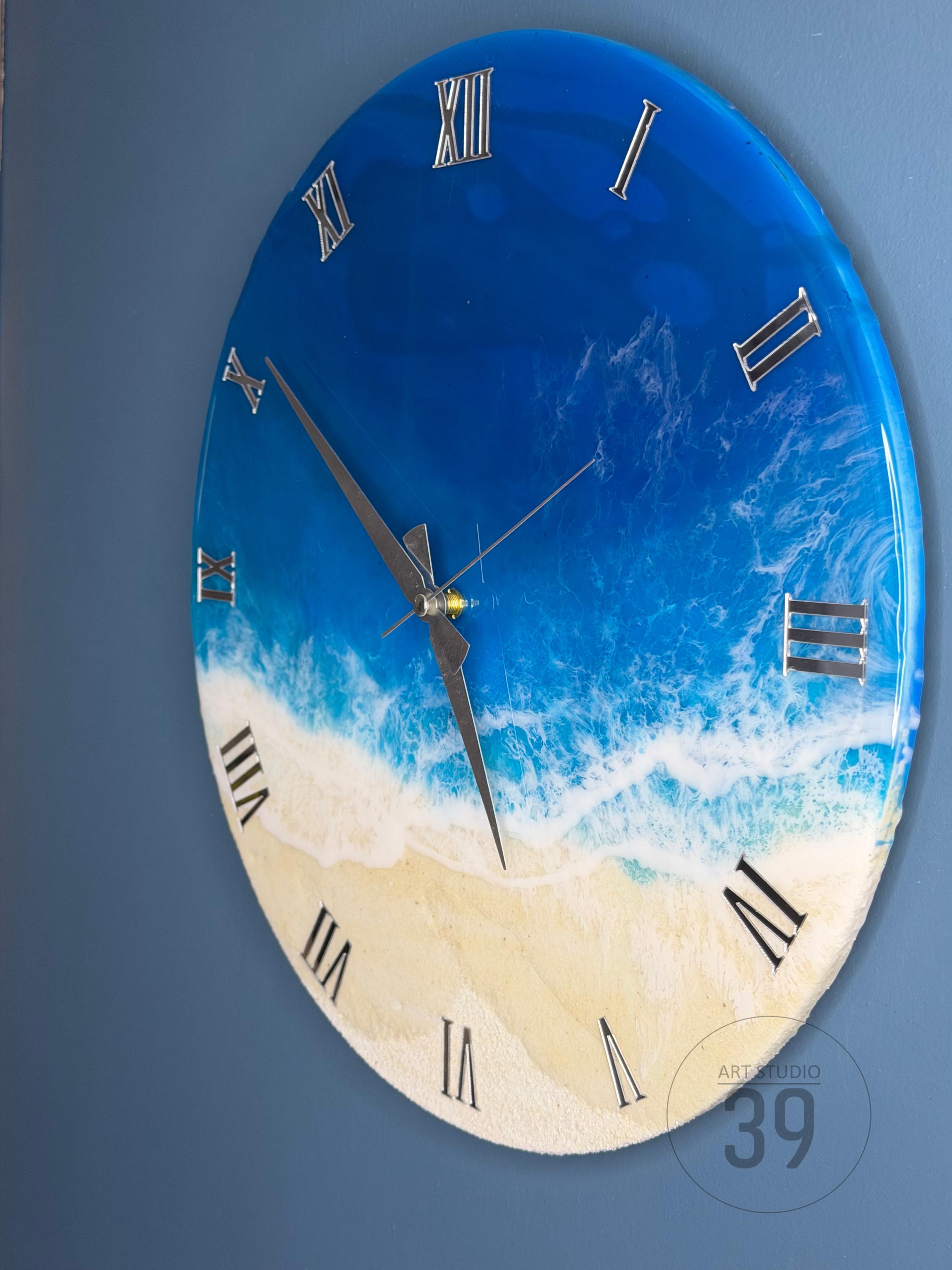 A round wall clock with a beach-themed design. The clock face features a gradient of blue and white, resembling the ocean, with a sandy beach at the bottom. Silver Roman numerals mark the hours. The clock hands are black.