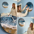 Round wall clock with a coastal design. The clock face features a depiction of ocean waves and a sandy beach, with Roman numeral hour markers. The clock hands are silver, and the clock is mounted on a light blue wall.