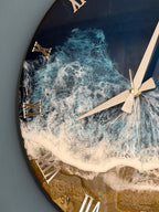 Round wall clock with a coastal theme. The clock face features a resin design depicting ocean waves in shades of blue and white, with a sandy beach at the bottom. Gold Roman numerals mark the hours, and silver hands indicate the time.
