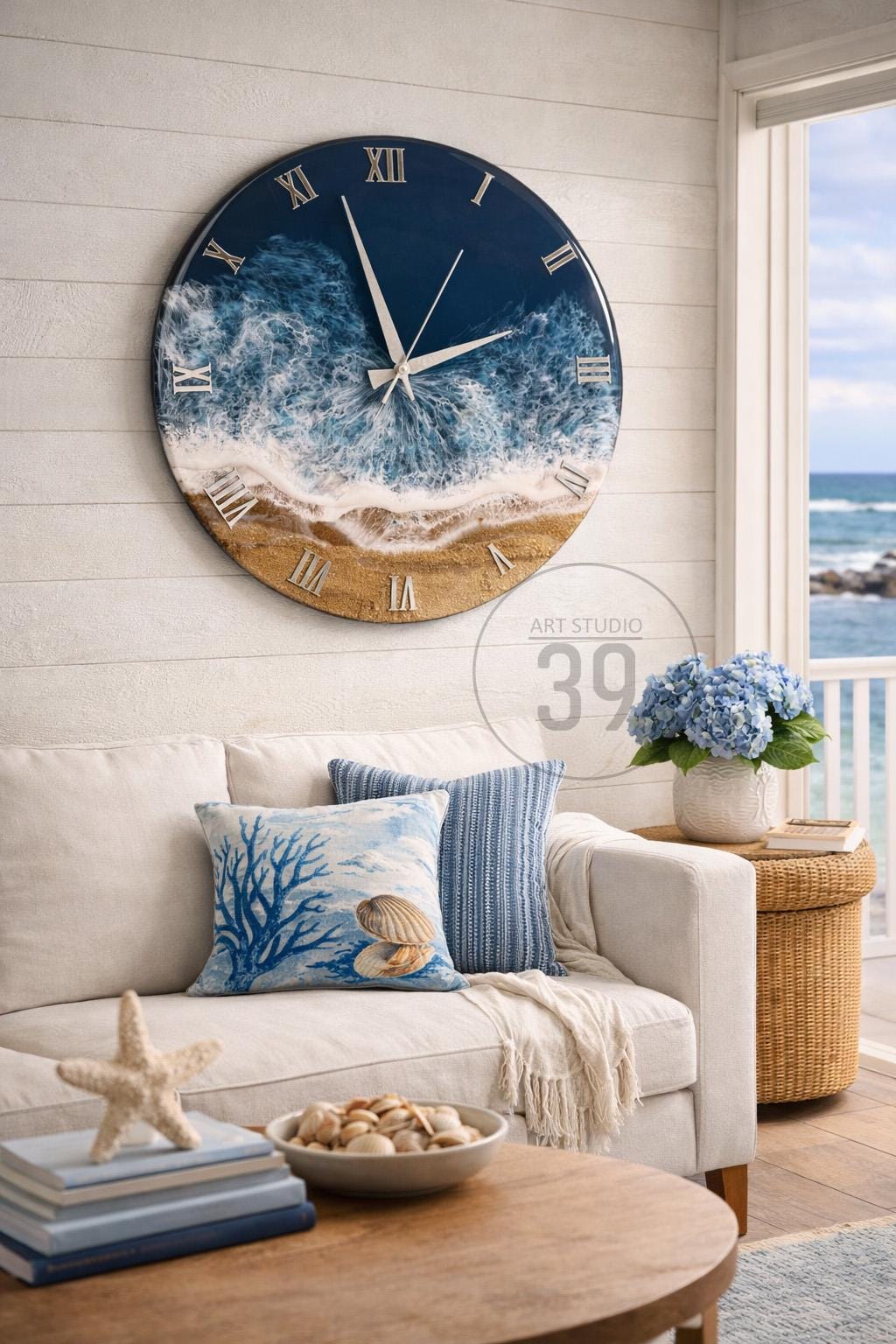 A large, round wall clock with a coastal theme. The clock face features a blue ocean design with white waves and a sandy beach. Silver Roman numerals mark the hours. The clock is mounted on a white wall.
