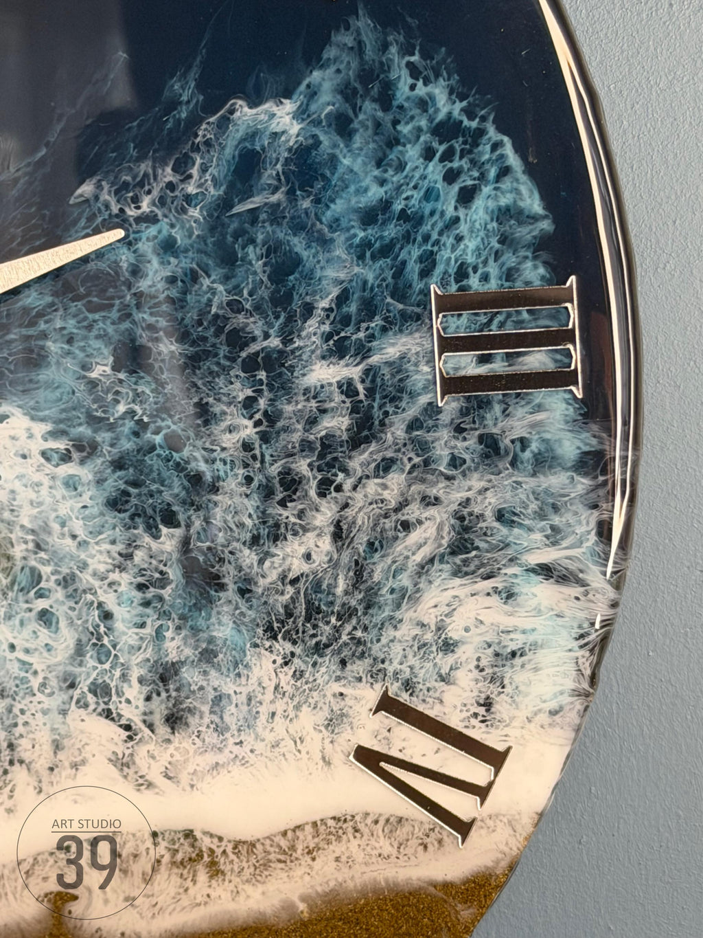 Round wall clock with a resin ocean wave design. The clock face features shades of blue and white, with Roman numerals and clock hands. The text "ART STUDIO 39" is visible on the bottom left.