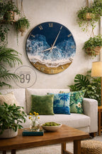 A large, round wall clock with a coastal theme. The clock face features a blue ocean design with white waves and a golden sand-like border. Roman numerals mark the hours. The clock is mounted on a light-colored wall.