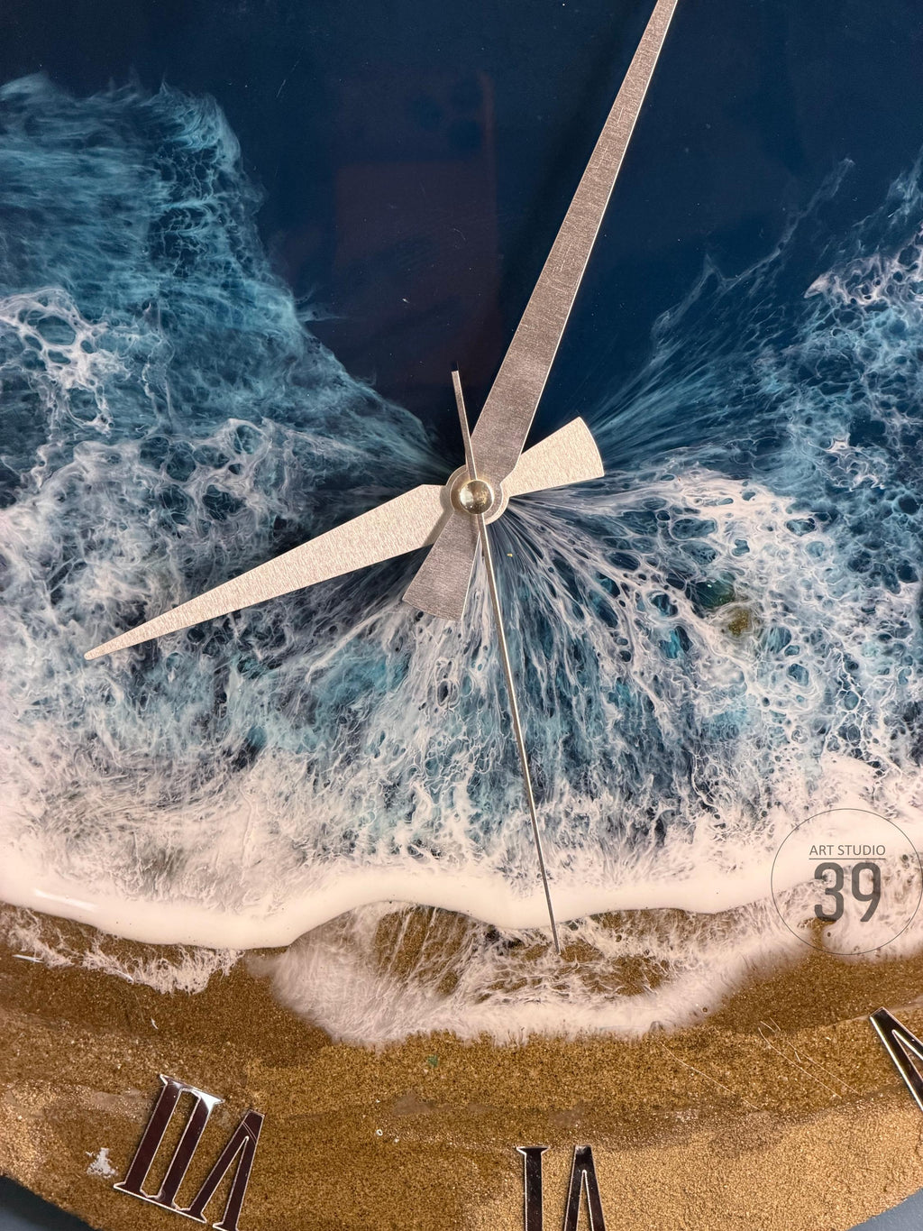 A round wall clock with a beach-themed design. The clock face features a depiction of ocean waves in shades of blue and white, with a sandy beach at the bottom. Silver clock hands and Roman numerals complete the design. The clock is labeled "ART STUDIO 39".