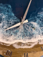 A round wall clock with a beach-themed design. The clock face features a depiction of ocean waves in shades of blue and white, with a sandy beach at the bottom. Silver clock hands and Roman numerals complete the design. The clock is labeled "ART STUDIO 39".