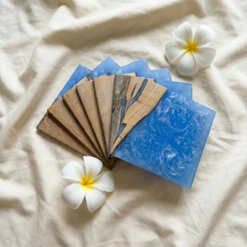 A set of handcrafted coasters featuring a combination of wood and blue resin. The coasters are square-shaped and arranged in a fan-like pattern. Two white plumeria flowers with yellow centers are placed near the coasters.