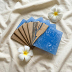 A set of handcrafted coasters featuring a combination of wood and blue resin. The coasters are square-shaped and arranged in a fan-like pattern. Two white plumeria flowers with yellow centers are placed near the coasters.